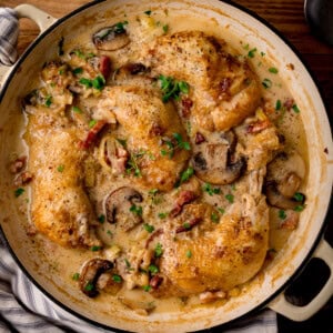 Overhead square image of a casserole pan filled with coq au vin blanc -cooked chicken breasts in creamy mushroom sauce. The pan is sitting on a striped napkin on a wooden table.