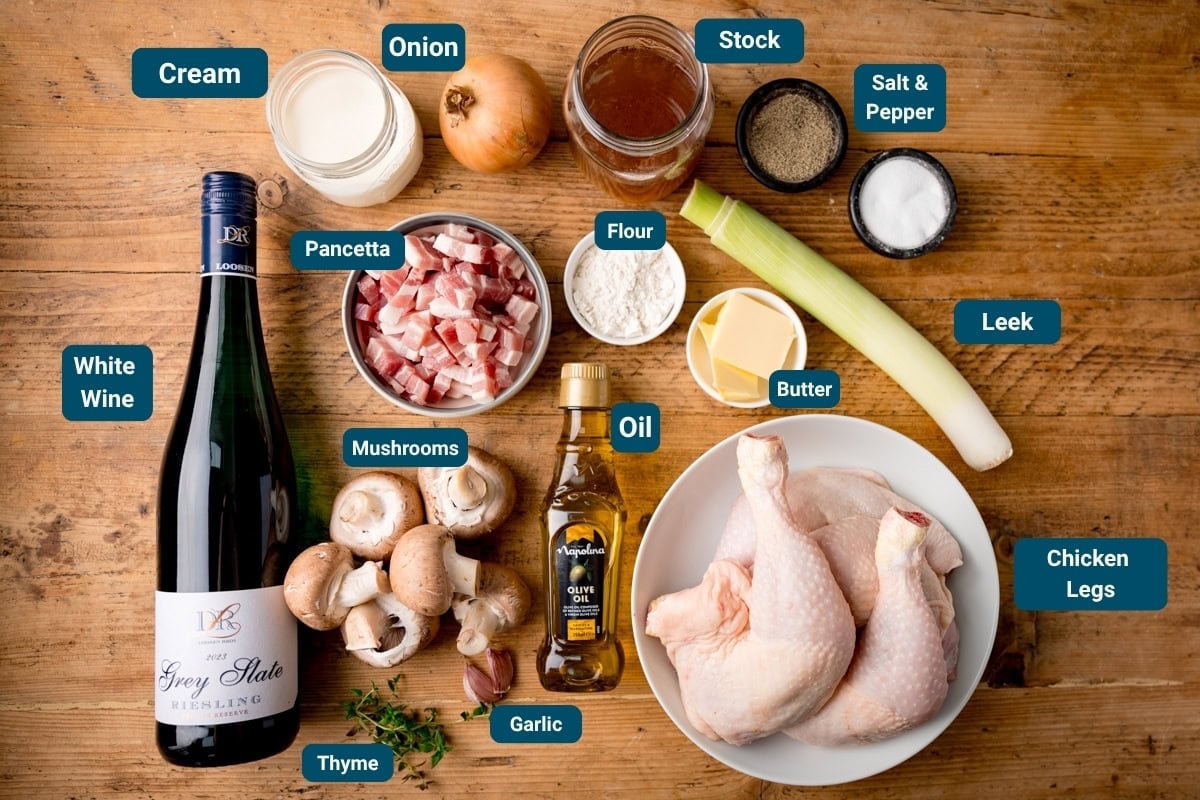 Ingredients for Coq au vin blanc on a wooden surface: chicken legs, leek, mushrooms, pancetta, cream, wine, stock, onion, garlic, oil, butter, flour, thyme, salt, and pepper.
