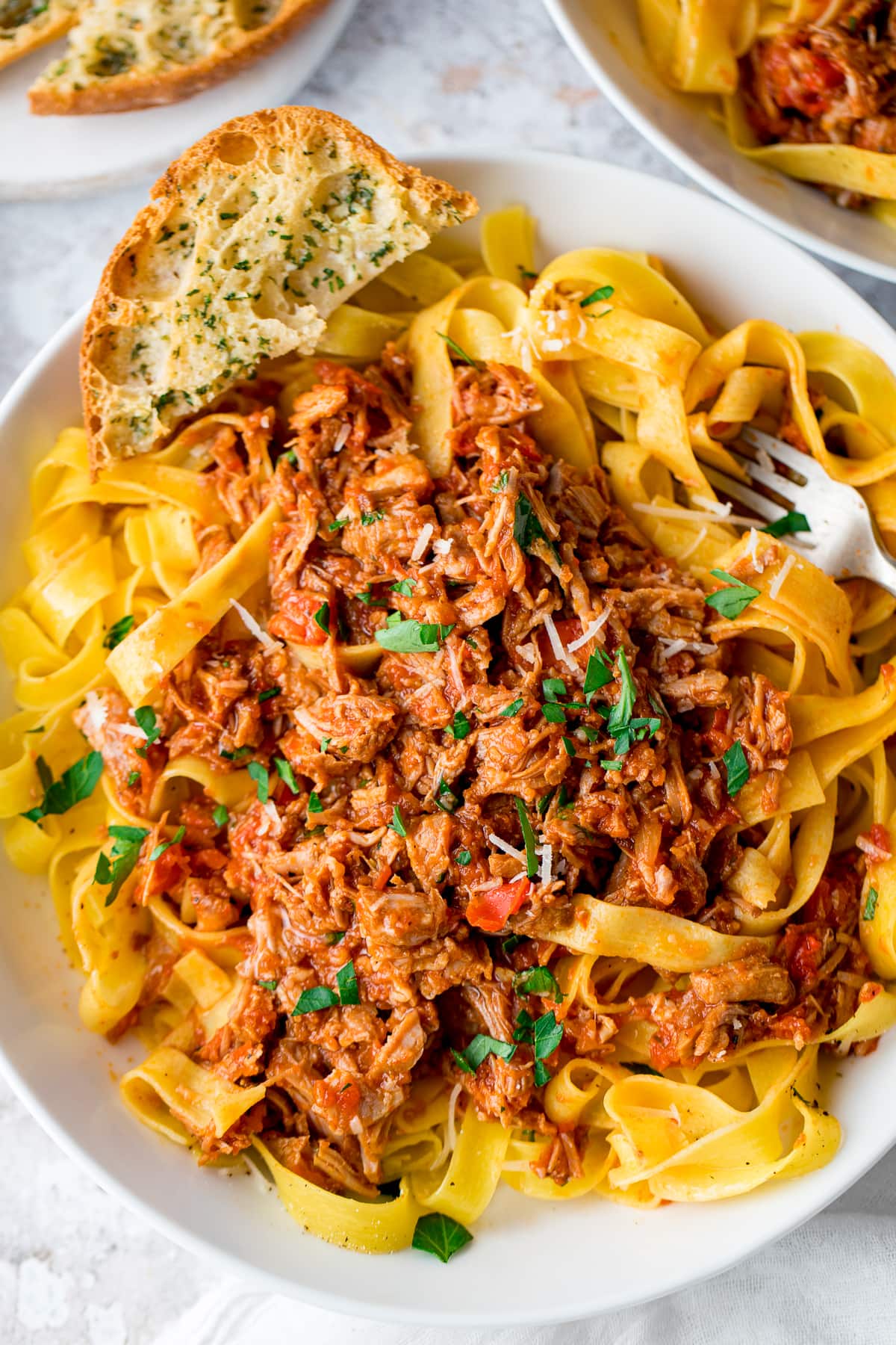 Overhead shot of slow cooked pork ragu with pasta in a white bowl with a slice of garlic bread