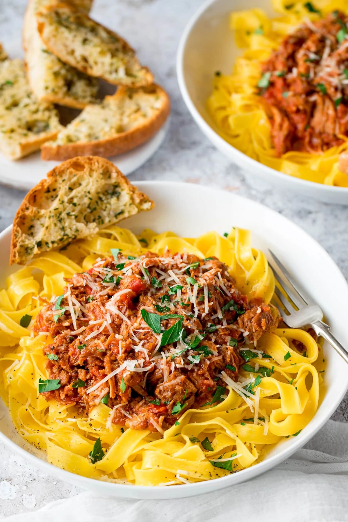 slow cooked pork ragu on top of pasta in a white bowl. Further bowl plus garlic bread in the background.