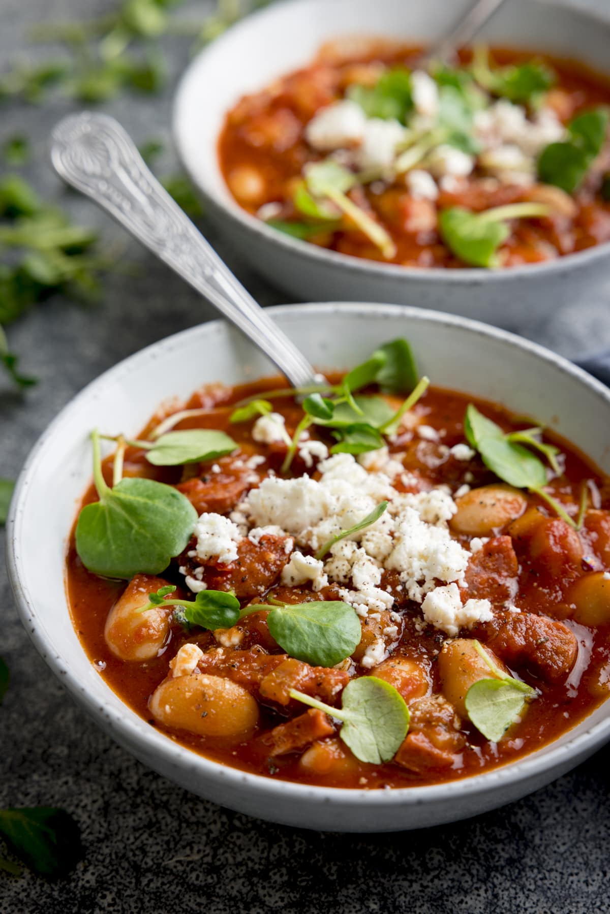Two light grey bowls filled with chorizo and butterbean soup, topped with feta and watercress. There is a spoon sticking out of the front bowl of soup.