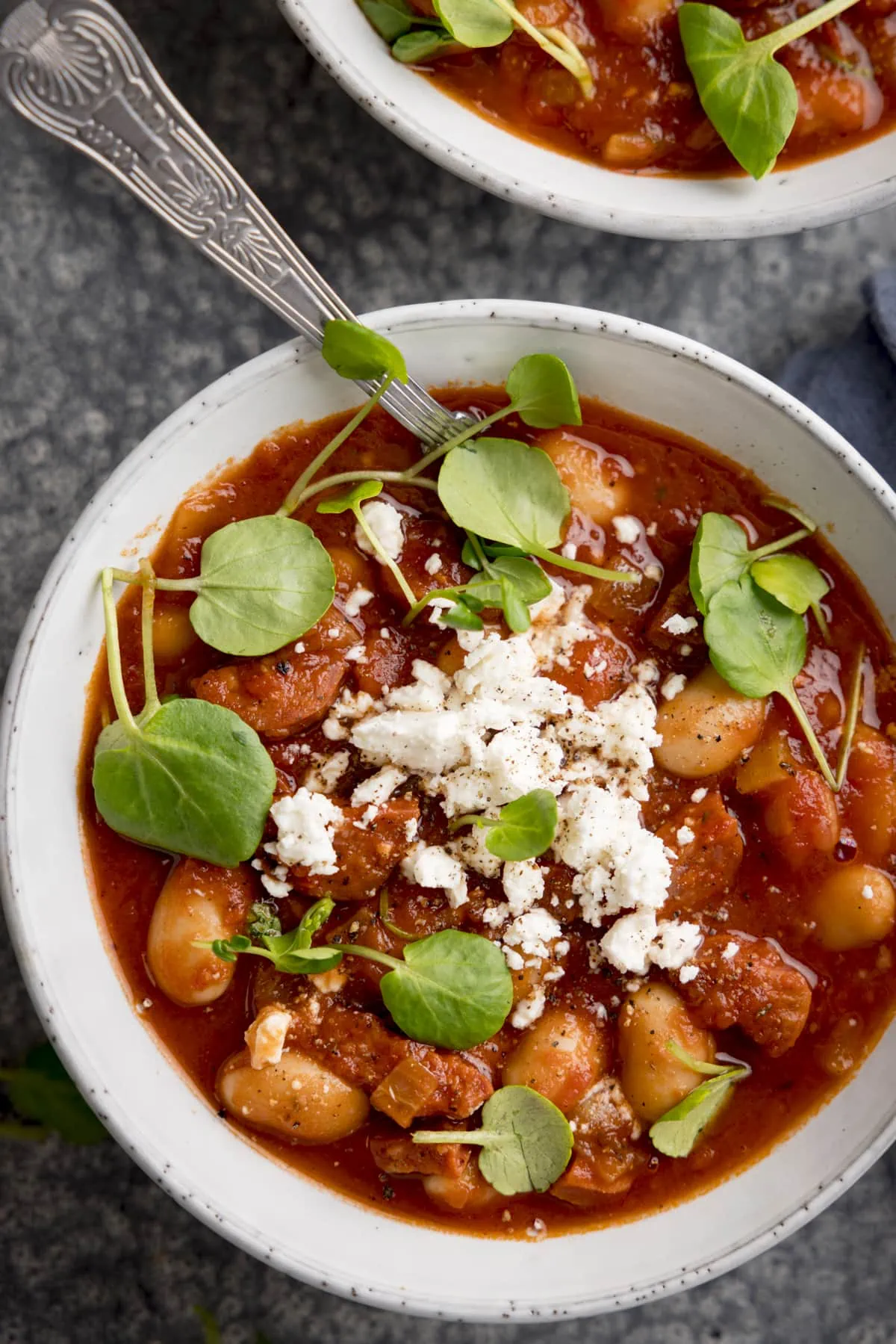 Overhead of butterbean and chorizo soup topped with feta and watercress in a light bowl. There is a second bowl just in shot at the top of the frame.