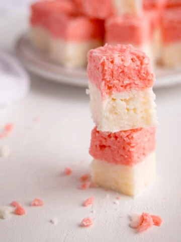 Two cubes of pink and white coconut ice stacked on top of each other on a white surface. There is more coconut ice in the background on a white plate, alongside a white napkin.