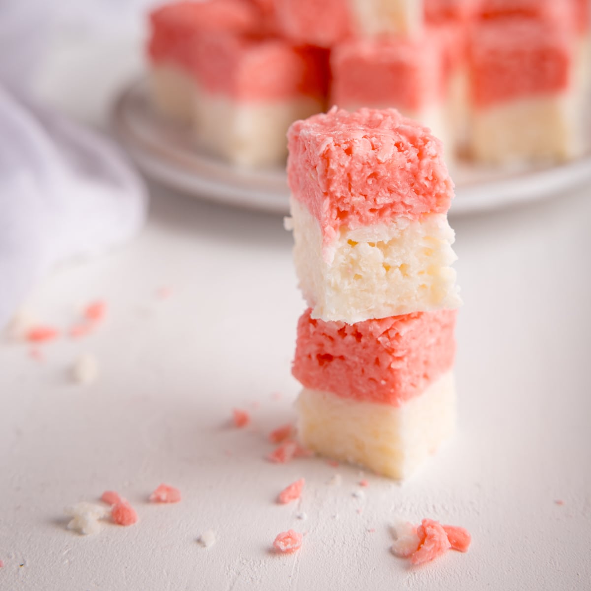 Two cubes of pink and white coconut ice stacked on top of each other on a white surface. There is more coconut ice in the background on a white plate, alongside a white napkin.