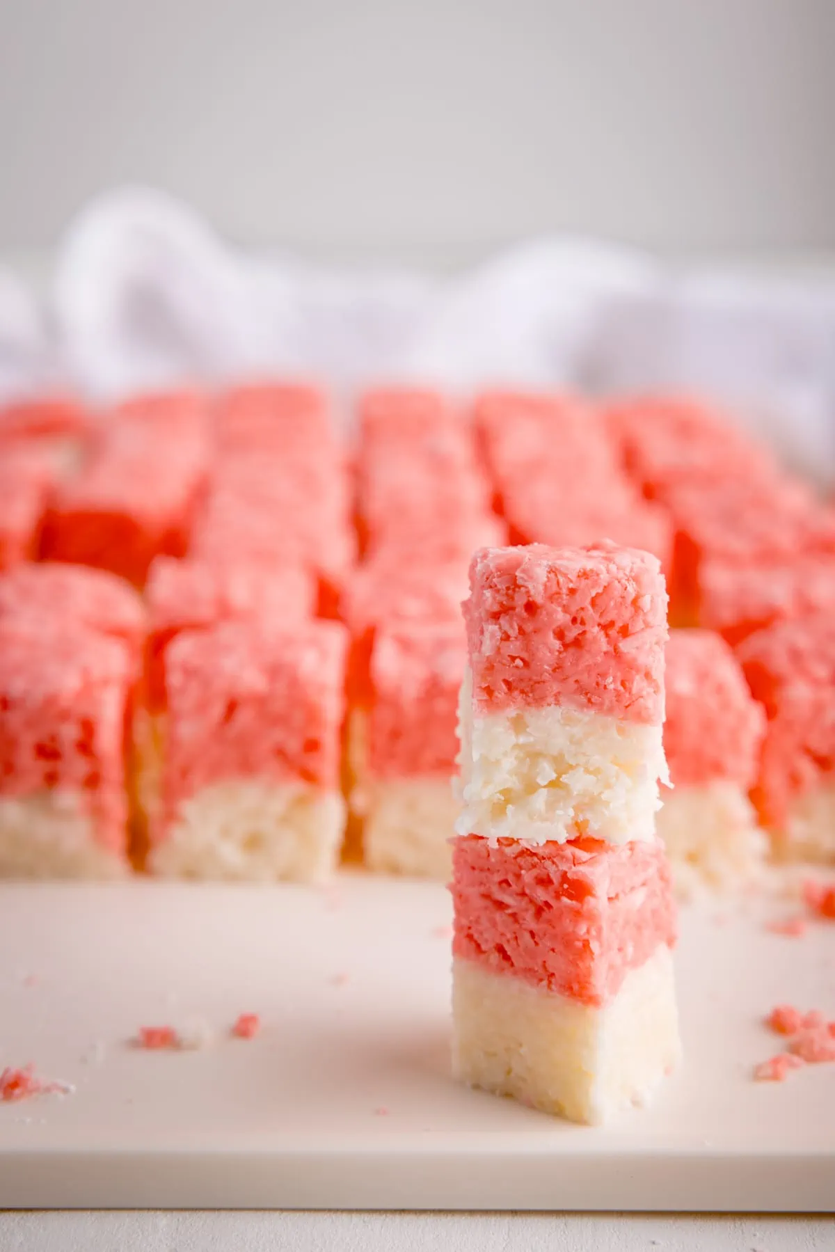 Pink and white coconut ice chopped into little cubes on a white board. There is a white napkin i the background.