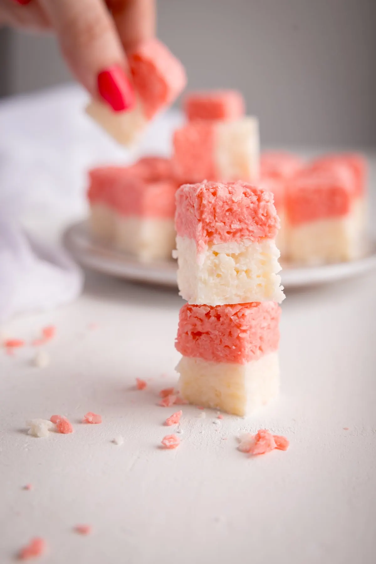 Two cubes of pink and white coconut ice stacked on top of each other on a white surface. There is more coconut ice in the background on a white plate, alongside a white napkin. One of the pieces of coconut ice in the background is being taking by a hand with pink-painted nails.