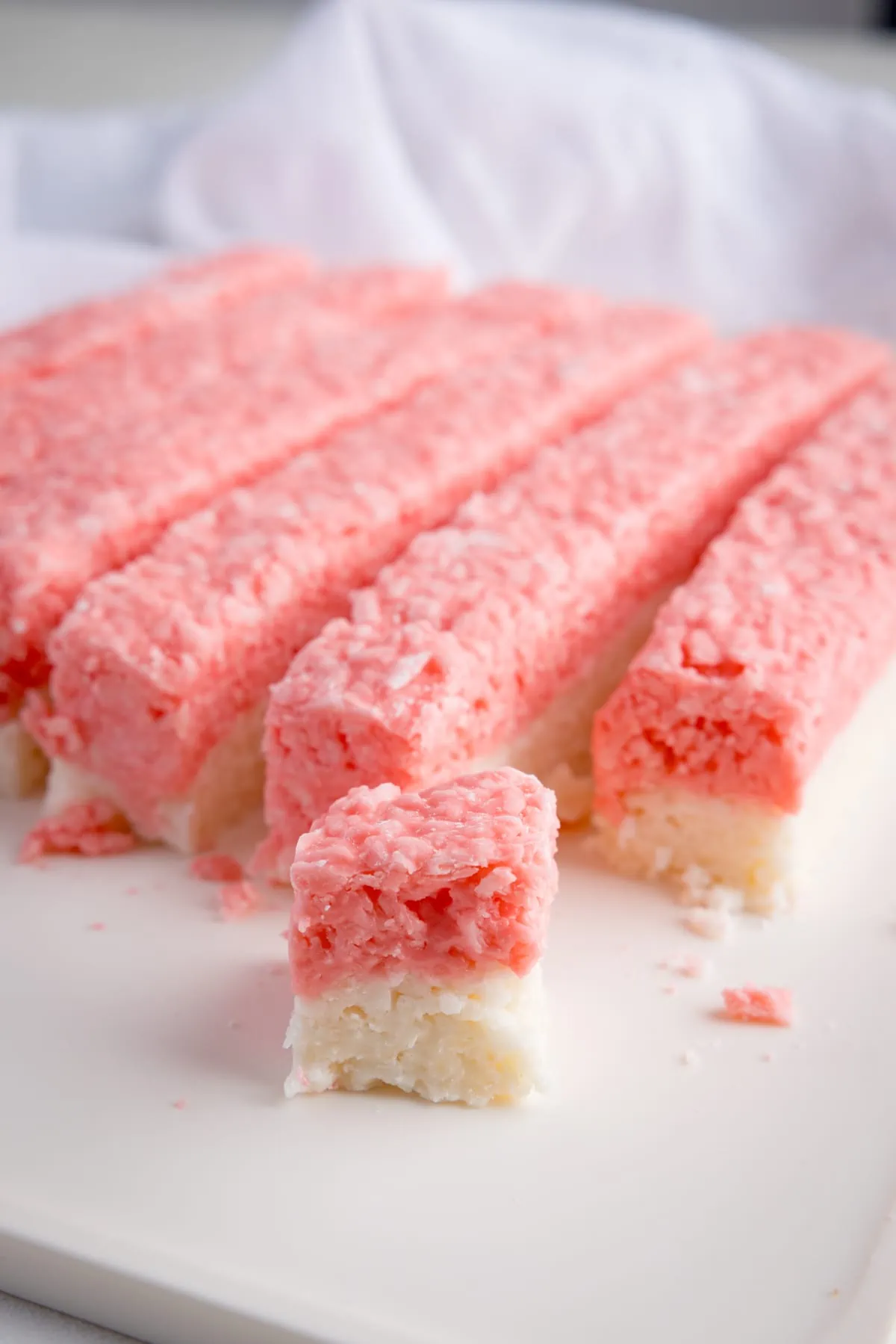 Pink and white coconut ice, sliced into long rows in a white board. There is one small cube of coconut ice in the foreground of the picture. There is a white napkin in the background.