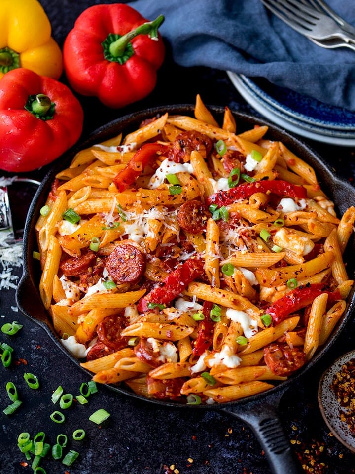Penne arrabiata pasta in a pan on dark background with peppers and spring onions scattered around pan