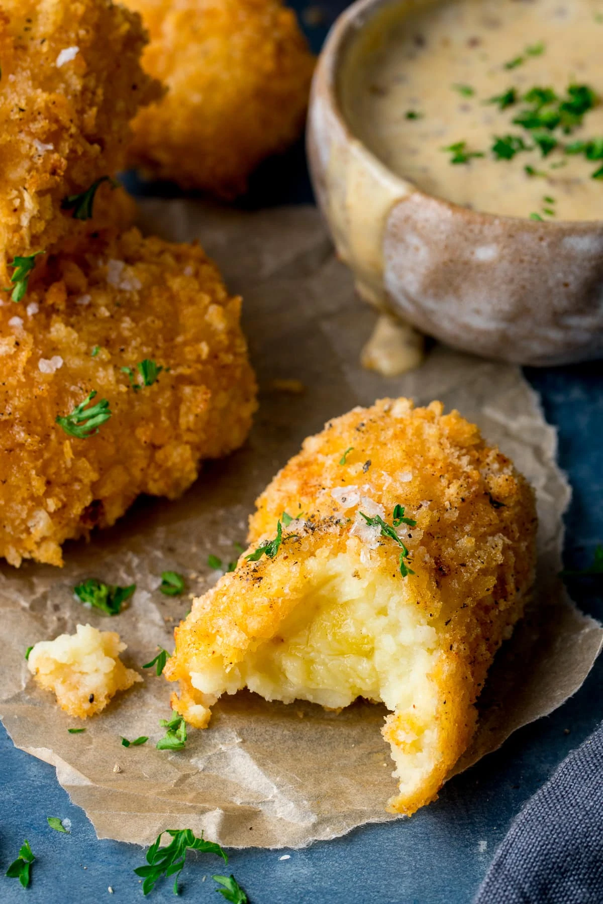 potato croquette with a bit taken out on a piece of parchment paper. Further potato croquettes and dip in the background.