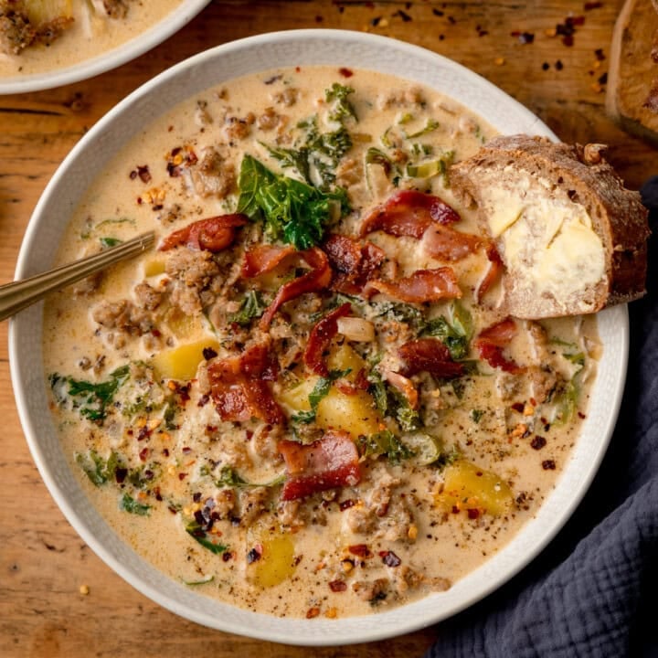 Zuppa Toscana Square FS A bowl of rich and creamy Zuppa Toscana with sausage, potatoes, kale, and bacon is served alongside a slice of buttered bread on a wooden table.