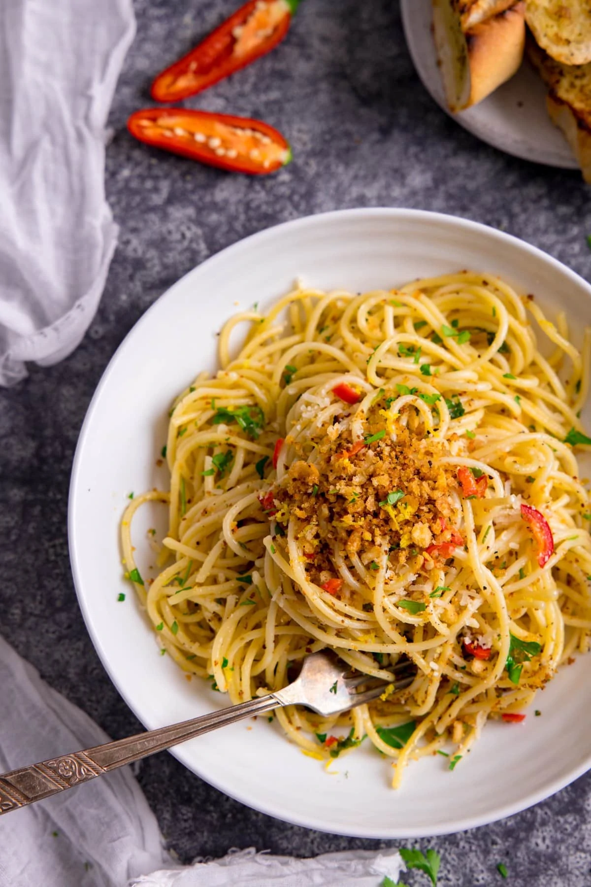 White bowl filled with garlic bread spaghetti. A fork is sticking out of the spaghetti. Bowl is on a grey background and there are ingredients and a white napkin scattered around.