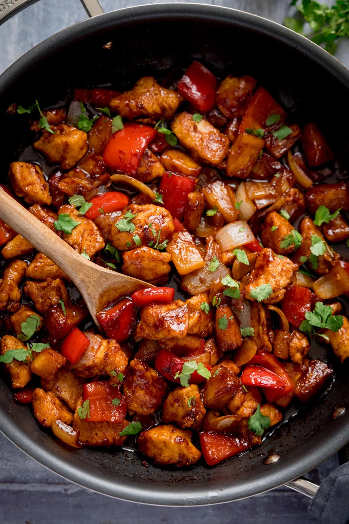 Tall overhead image of Sticky Pineapple Chicken Stir Fry in a pan. There is a wooden spoon sticking out of the pan.