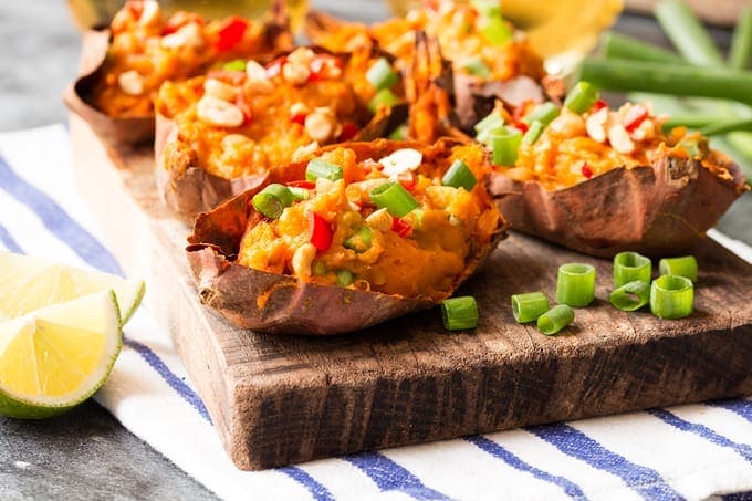 Thai inspired sweet potato skins on a wooden board on top of a striped tea towel. The potato skins are topped with spring onions, chillies and peanuts. There are additional ingredients scattered around.