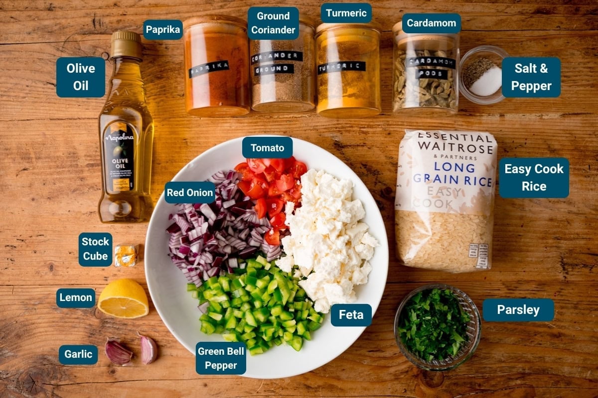 Overhead view of labelled ingredients for vegetable and feta rice including: chopped vegetables, spices, feta, olive oil, rice, garlic, lemon, stock cube and parsley. All the ingredients are labelled and they sit on a wooden surface.