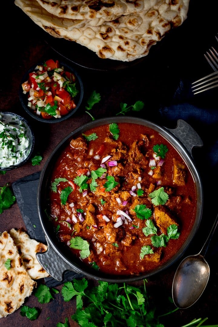Overhead of slow cooked beef curry in a dish on a dark background with ingredients scattered around