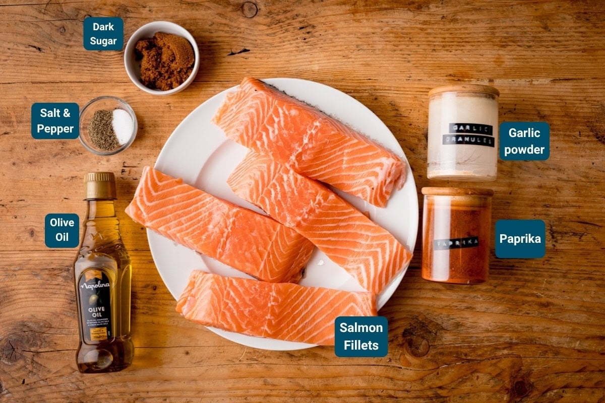 A plate of raw salmon fillets for on a wooden table, surrounded by olive oil, salt and pepper, dark sugar, garlic granules, and paprika. Each ingredient for this Salmon recipe is clearly labelled.