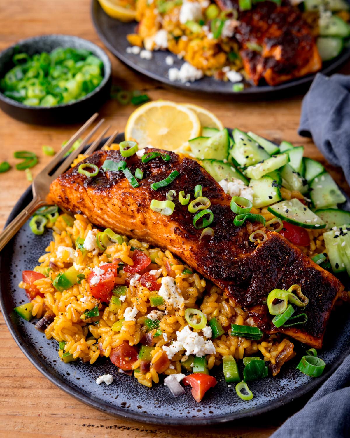 A plate with spiced pan-fried salmon on top of yellow rice with vegetables and feta cheese, cucumber and lemon slices, topped with chopped scallions. There is a further plate and a small dish of scallions in the background.