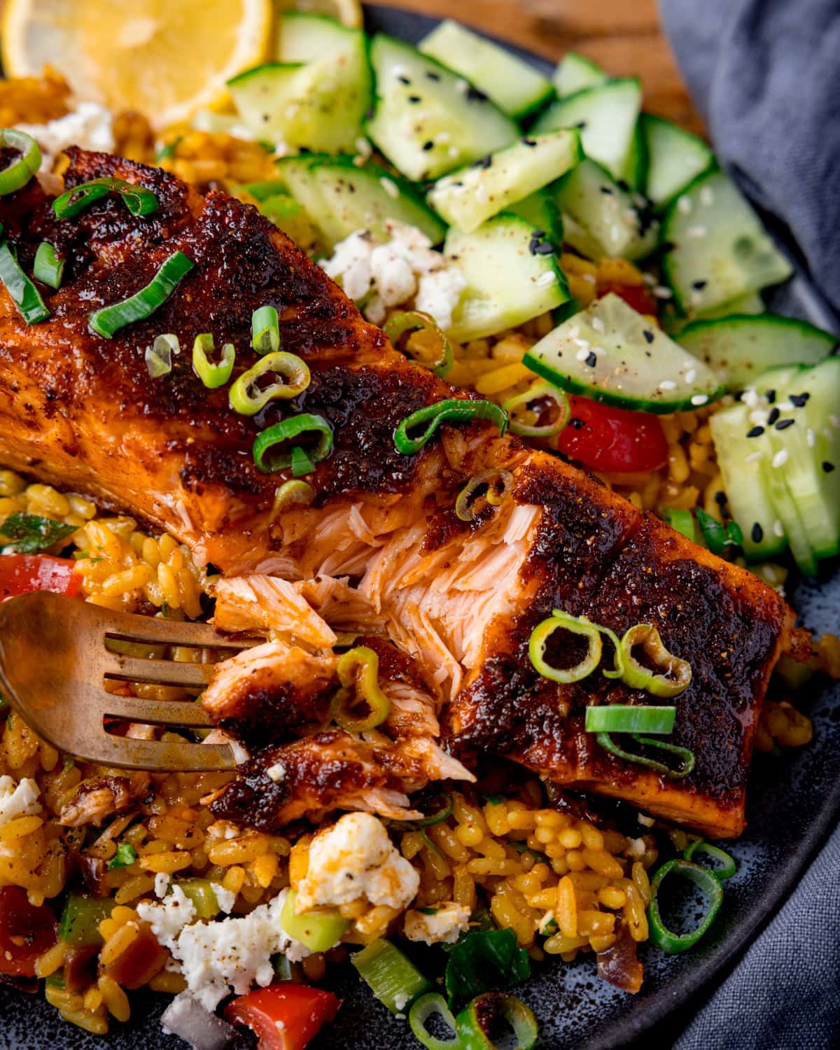 A close up of a plate of pan-fried salmon served on spiced rice with chopped vegetables, feta cheese, cucumber slices, and a lemon wedge, garnished with spring onions. A fork is taking a piece of the salmon.