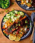 A dark plate with spicy salmon fillet on top of vegetable and feta rice with sliced cucumbers, scallions, sesame seeds, and lemon slices, served with a fork and knife on the side. There is a further plate partially in shot at the top of the image and also a small bowl of chopped scallions.