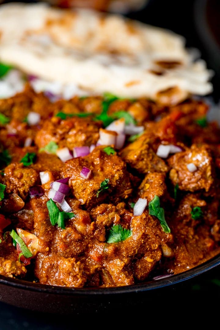 Close up pf slow cooked beef curry with coriander and red onion on top. Torn pitta bread in background.