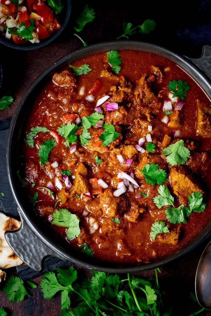 Overhead image of beef curry in a dark dash on a dark background, topped with coriander, chillies and red onion