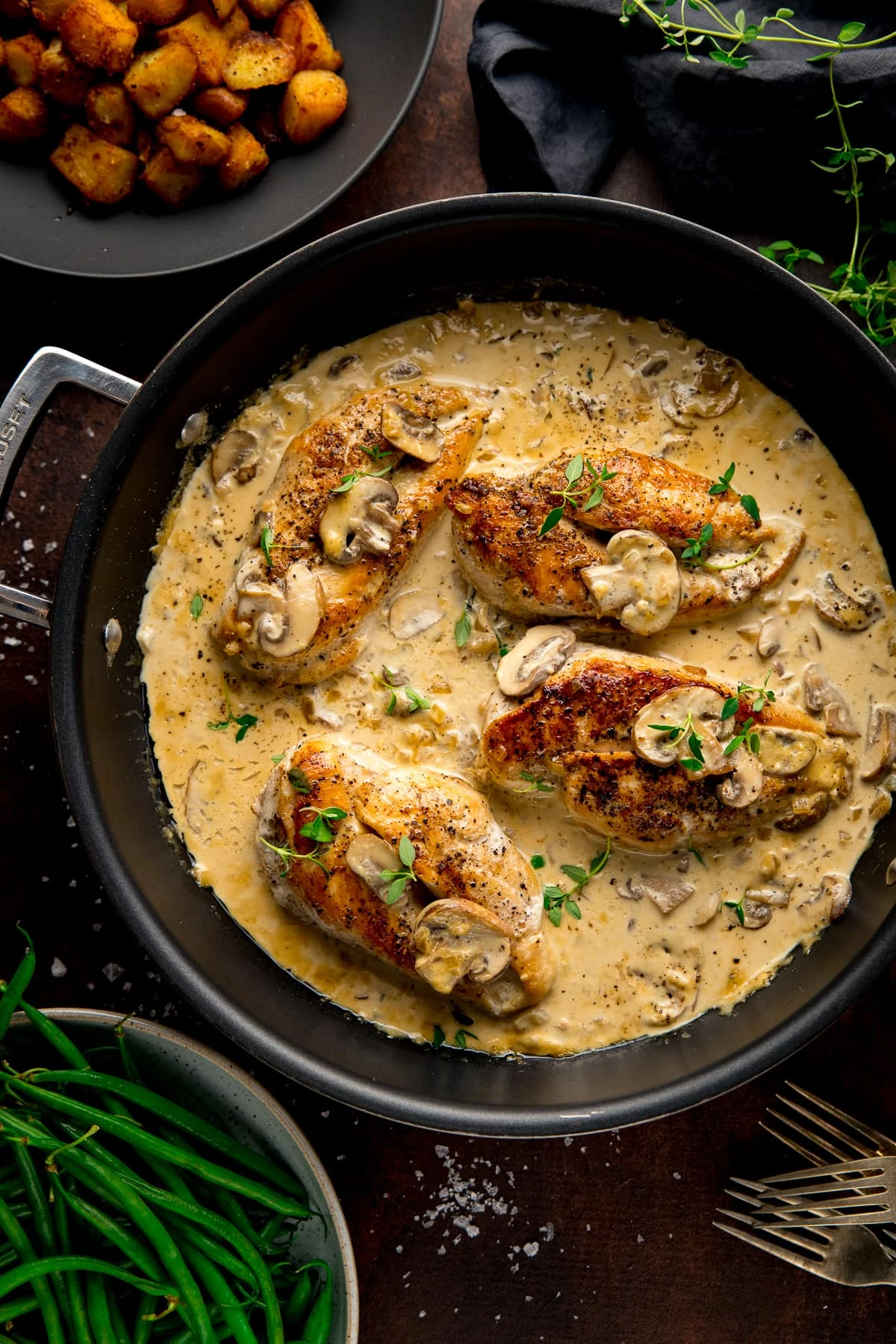 Pan with chicken in creamy white wine sauce and mushrooms, next to a bowl of green beans and a bowl of saute potatoes