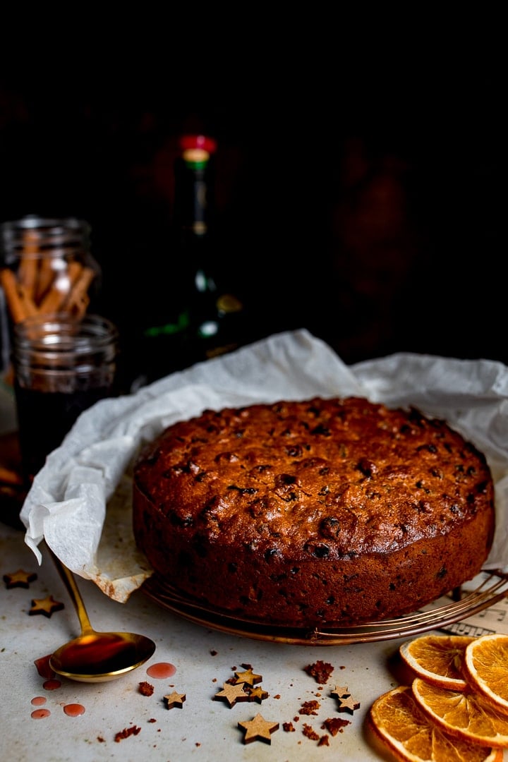 A fruit cake sat on a cooling rack with a spoon of cherry brandy to one side and some festive Christmas decorations in the foreground.