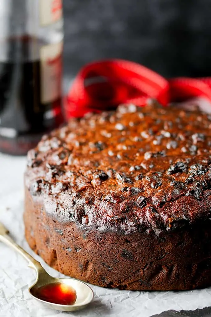 A fruit cake sat on a cooling rack with a spoon of cherry brandy to one side and some festive Christmas decorations in the foreground.