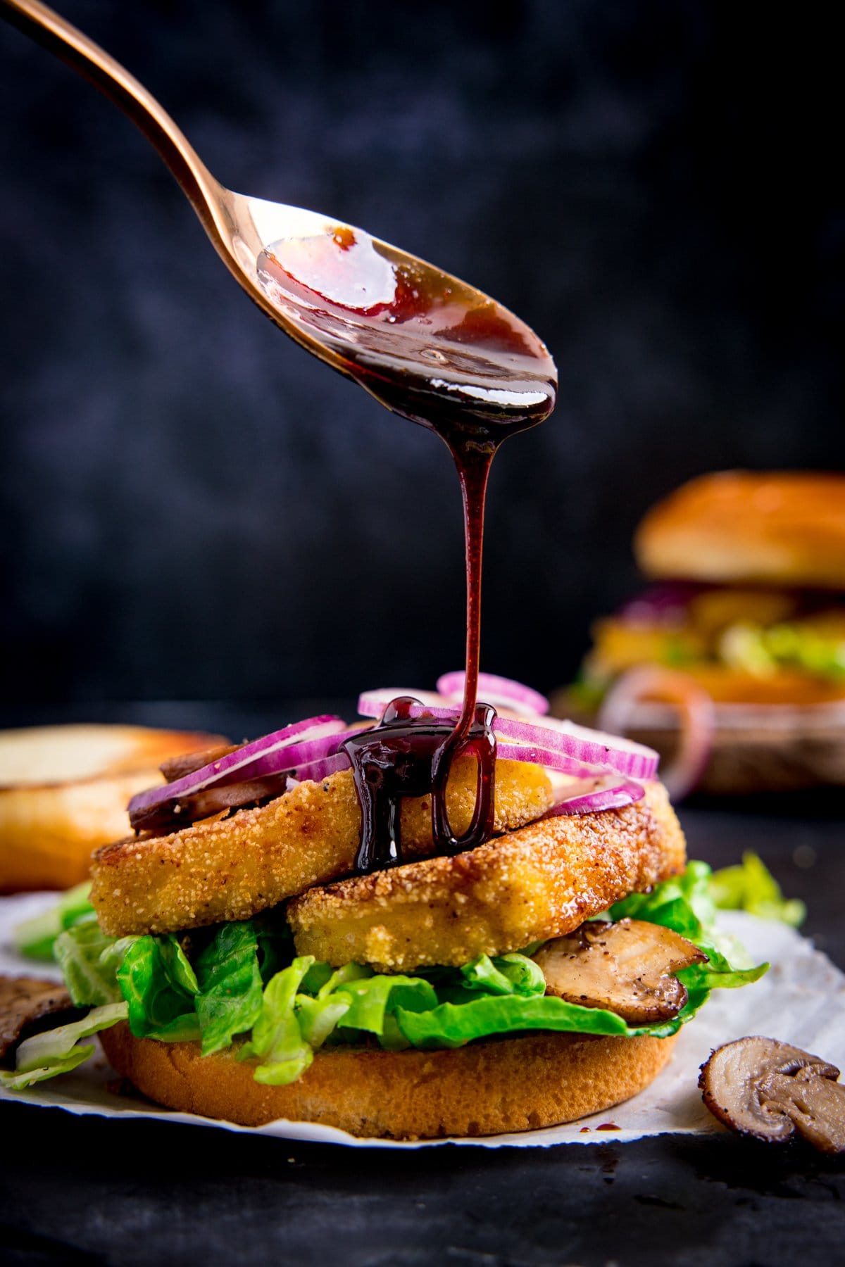 Sticky chilli glaze being drizzled from a spoon onto an open crispy halloumi burger. The burger is on a piece of parchment against a dark background.