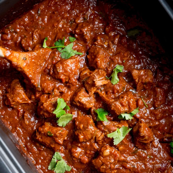 Square close-up overhead image of slow cooker pork and beef chilli in a slow cooker with a wooden spoon sticking out. There is a little sprinkle fresh coriander on top.