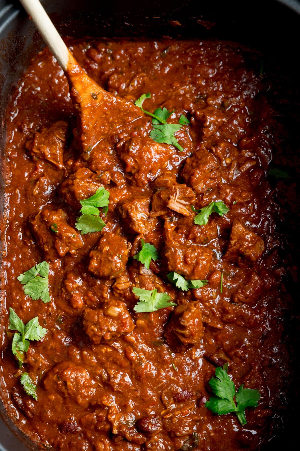 Close-up overhead image of slow cooker pork and beef chilli in a slow cooker with a wooden spoon sticking out. There is a little sprinkle fresh coriander on top.