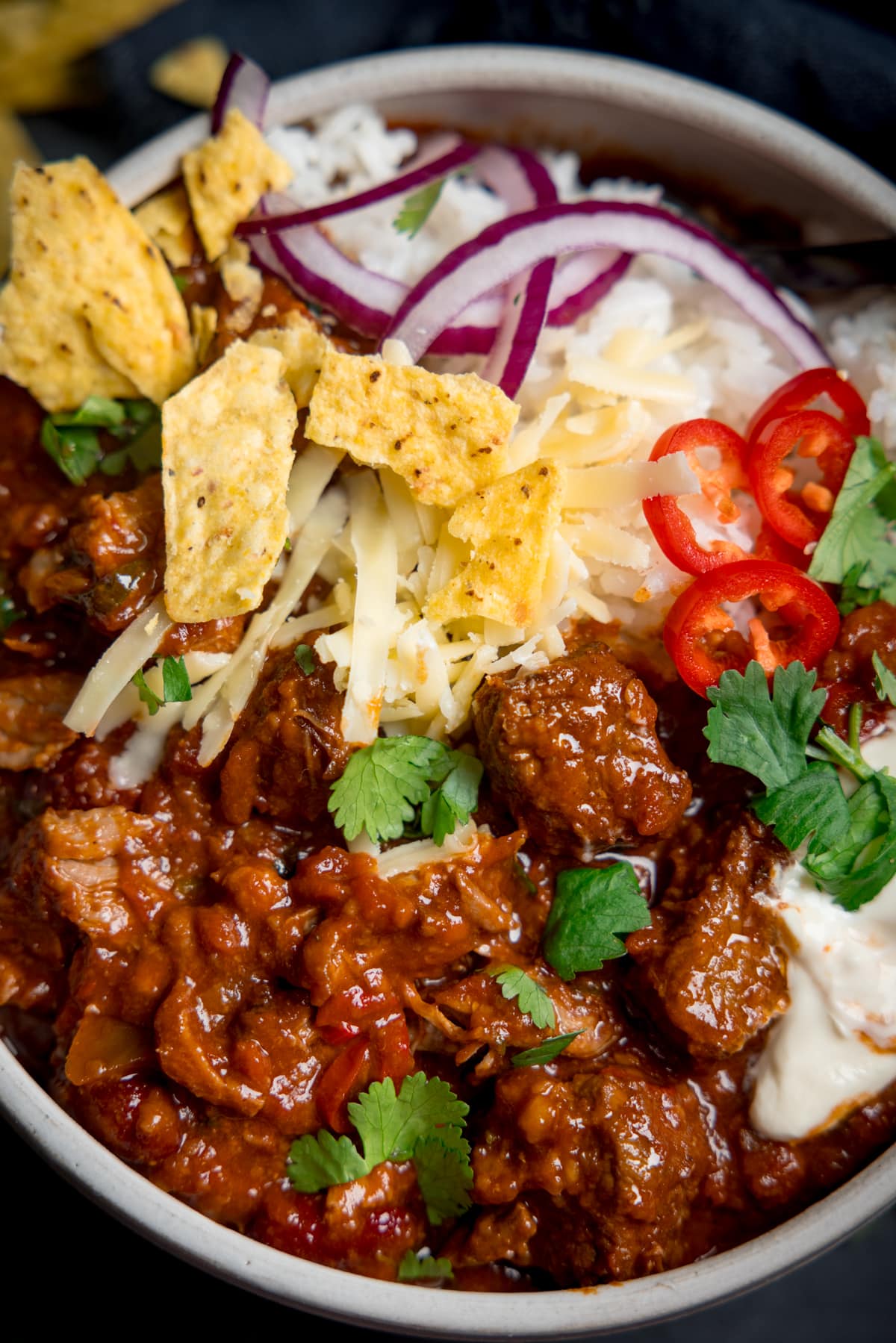 Overhead of pork and beef chilli in bowl with rice, sour cream, sliced red chillies, grated cheese, sliced red onion, fresh coriander and crumbled tortilla chips on top.