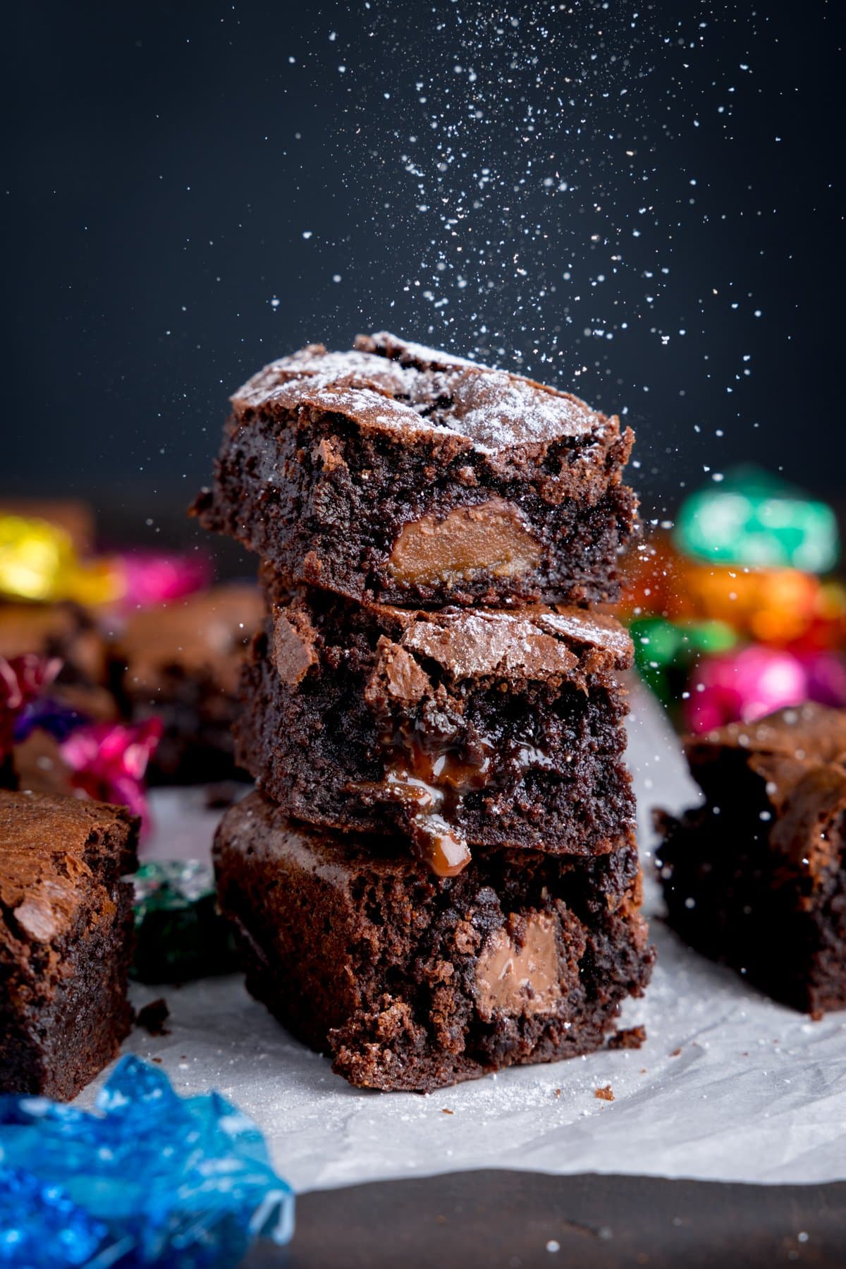 A pile of chocolate Brownies on some baking parchment with icing sugar being dusted on with some empty chocolate wrappers in the background.