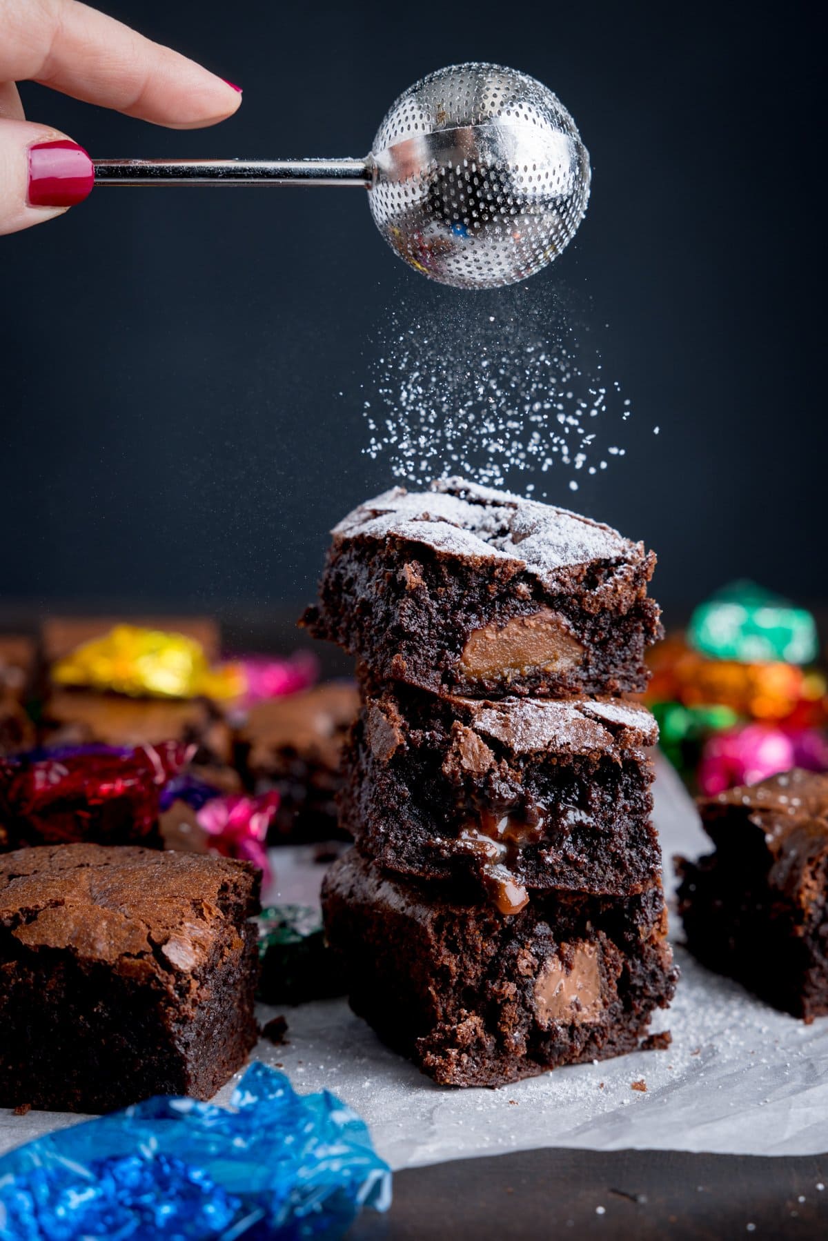 A pile of chocolate box brownies on some baking parchment with icing sugar being dusted on from a mini sieve with some empty chocolate wrappers in the background.
