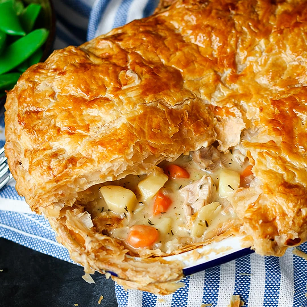 Side on angled close up photo Creamy Chicken and Vegetable puff pie with a slice taken out showing the carrots, chicken potato and creamy sauce inside. Dish is sat on a blue and white striped cloth with green veg in the background