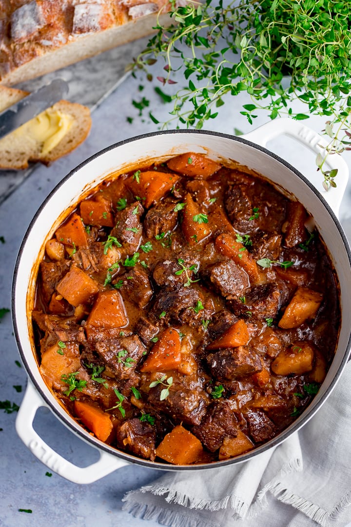 Large white pan filled with Scottish beef stew on a light background. Bread and fresh herbs also in scene.