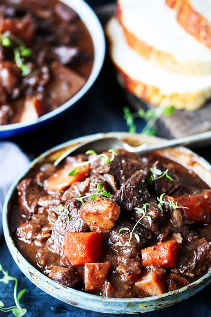 Large white pan filled with Scottish beef stew on a light background. Bread and fresh herbs also in scene.