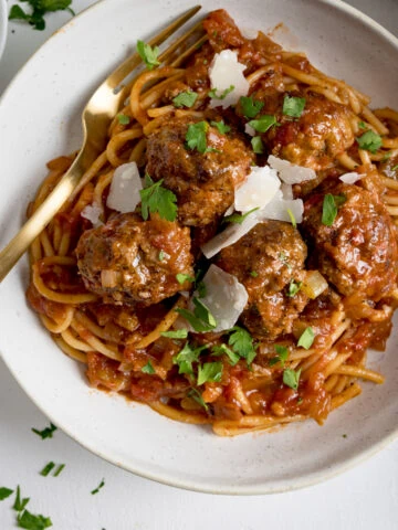 Spaghetti and meatballs in tomato sauce in a white bowl on a white background. There is a gold fork in the bowl.