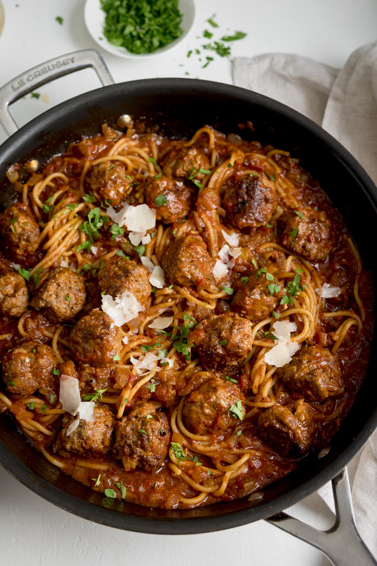 Overhead image of spaghetti and meatballs in a large frying pan on a white background. There is a small bowl of chopped parsley at the top of the image.