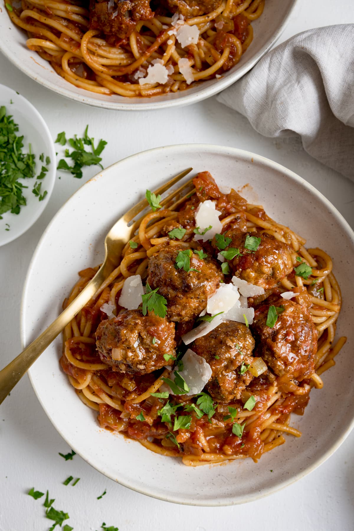 Spaghetti and meatballs in tomato sauce in a white bowl on a white background.  There is a golden fork in the bowl.  There's another bowl of spaghetti and meatballs right at the top of the frame.