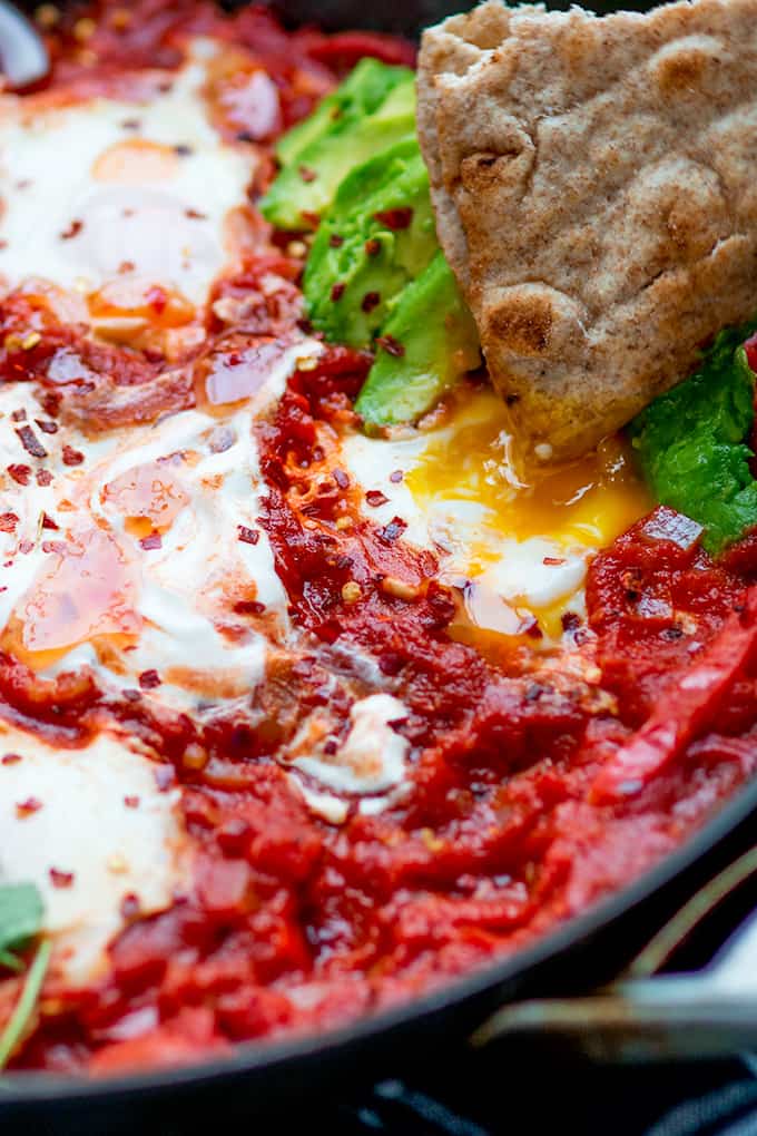 Close up shot of Mexican shakshuka in a pan with avocado and chilli flakes. A piece of flatbread is being dipped in an egg yolk.