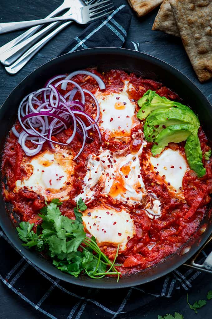 Overhead image of Mexican shakshuka in a pan with avocado, red onion, coriander and chilli flakes. The pan is on a dark background with flatbreads and forks just in shot.