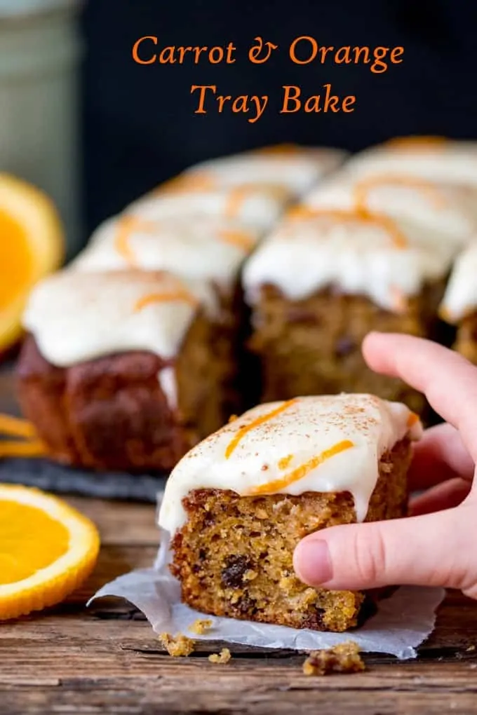 a hand grabbing a piece of carrot tray bake with orange infused raisins and zesty cream cheese frosting