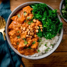 A bowl of rice topped with a Chicken Sweet Potato and Peanut Stew, featuring peanuts and chopped greens, garnished with fresh herbs. It sits on a wooden table, accompanied by a fork and teal napkin.