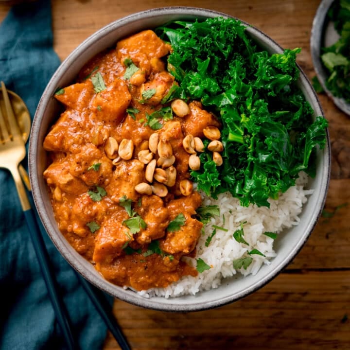 A bowl of rice topped with a Chicken Sweet Potato and Peanut Stew, featuring peanuts and chopped greens, garnished with fresh herbs. It sits on a wooden table, accompanied by a fork and teal napkin.