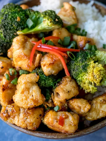 Square image of Asian crispy chicken with broccoli and rice in a stoneware bowl. Blue napkin in background