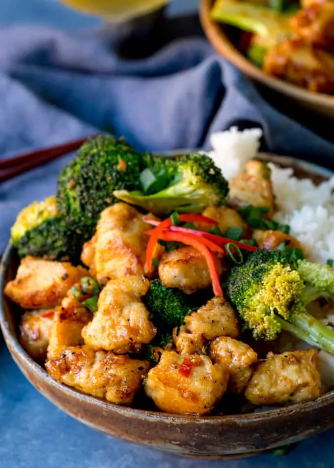 Side on shot of crispy chicken with broccoli and rice in a bowl with blue background. Further bowl in backround.