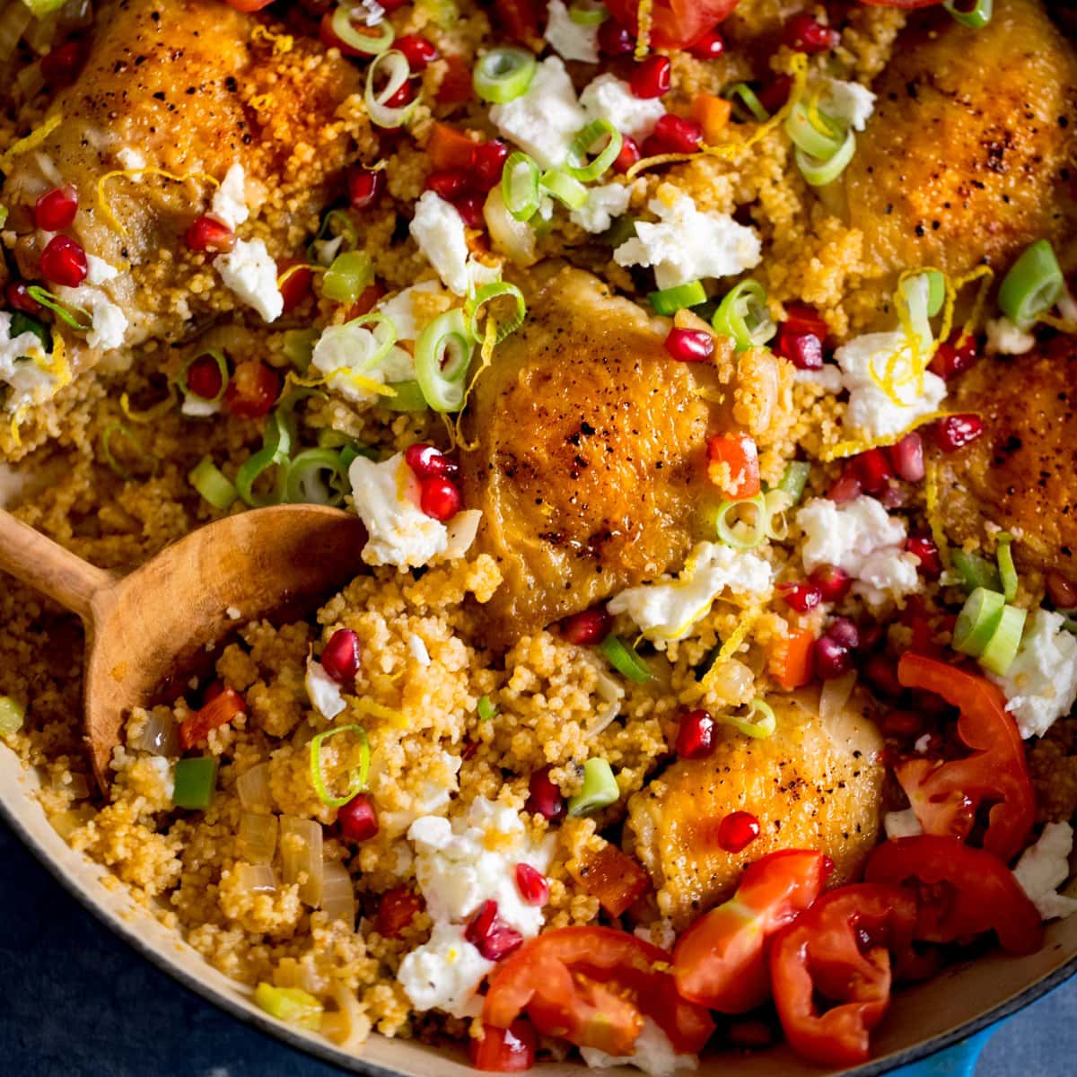 Square image of one-pot chicken and couscous in a blue pan, topped with feta and pomegranate. There is a wooden spoon digging into the couscous.