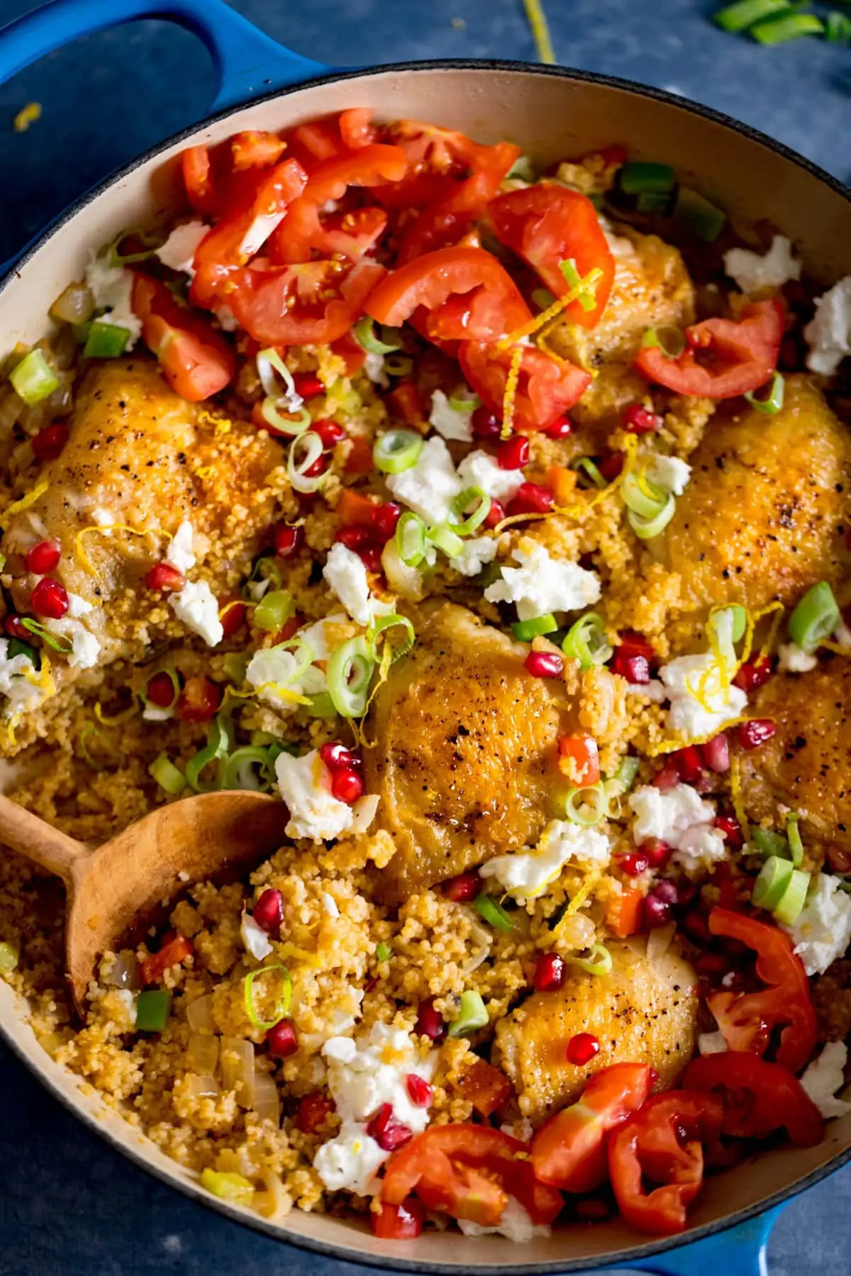 Chicken and couscous one pot in a pan on a blue table. There is a spoonful being taken from the pan. The dish is topped with feta, pomegranate and chopped tomatoes.