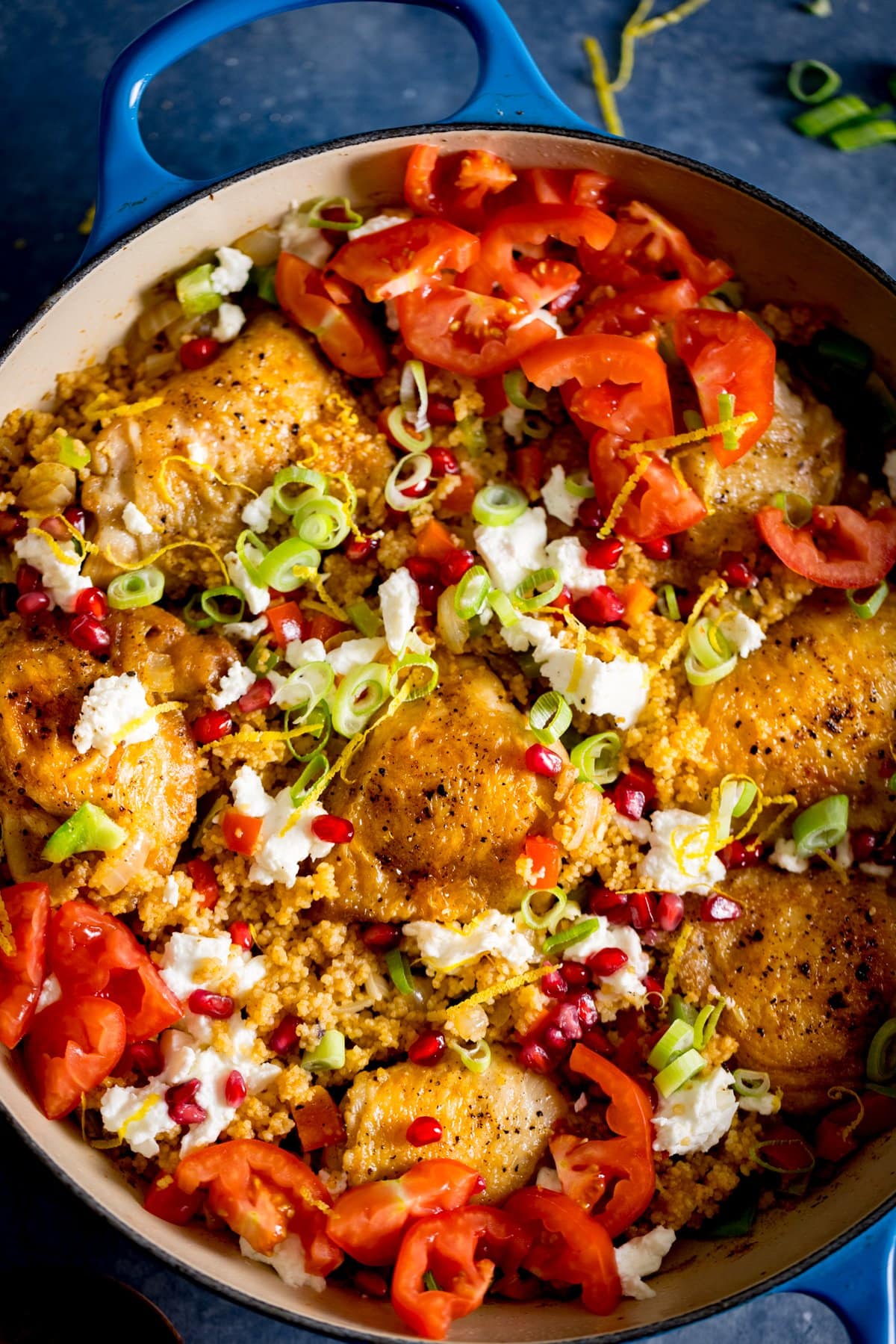 Overhead image of one-pot chicken and couscous in a blue pan, on a blue table, topped with feta, tomatoes and pomegranate.